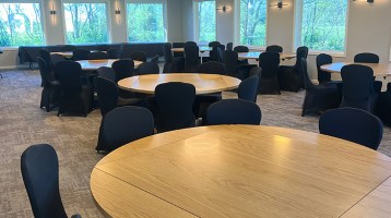 A modern, well-lit event or conference room with multiple round wooden tables surrounded by black upholstered chairs, large windows letting in natural light, and contemporary ceiling and wall lighting.
